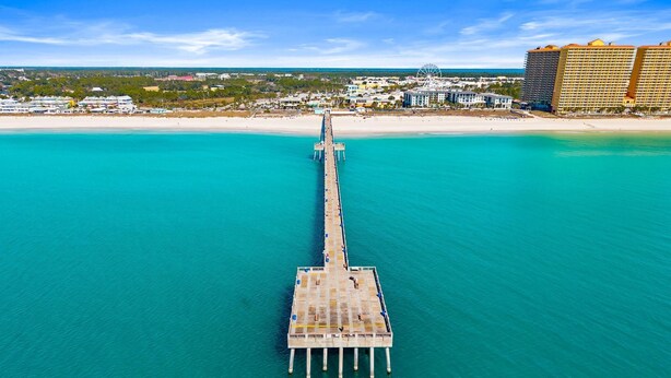 Luftaufnahme eines Piers, der sich in türkisblaues Wasser erstreckt, mit einem Strand und Hotelgebäuden im Hintergrund.