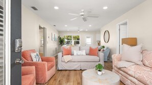Ein großzügiges Wohnzimmer mit grauem Sofa, einem rosa Sofa und einem runden Couchtisch. Eine Deckenleuchte, eine Tür mit Zahlenschloss und ein Fenster mit Blick auf Grünflächen sind ebenfalls vorhanden.