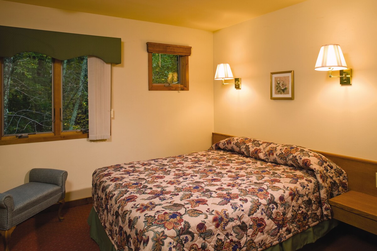 A bedroom with a floral bedspread, a bench, two wall-mounted lamps, and a window with a view of trees.