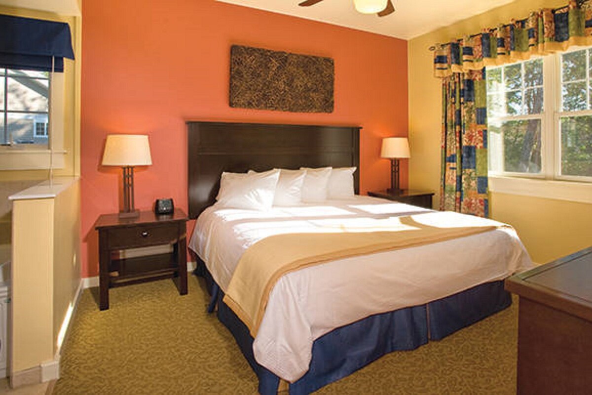 A bedroom with a large bed, two bedside tables with lamps, a headboard, a window with curtains, and a wall-mounted artwork.