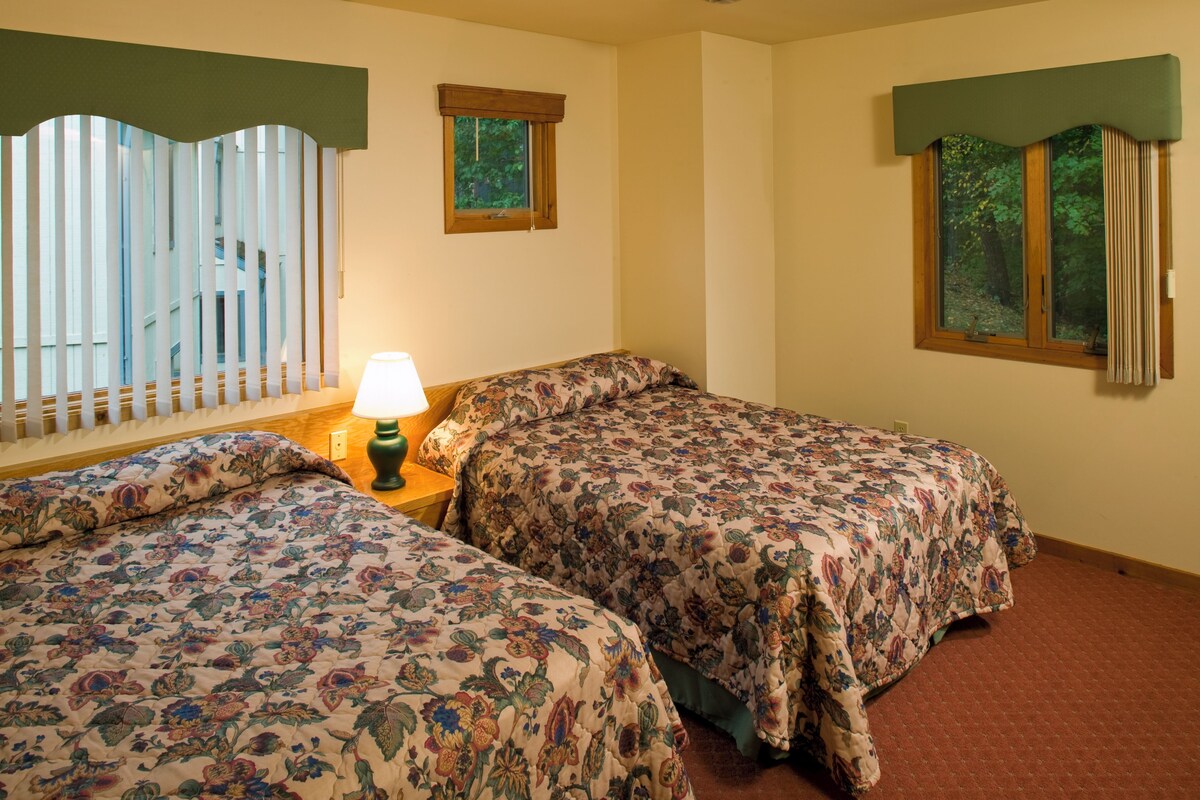 A room with two beds, a nightstand with a lamp, and two windows with curtains.