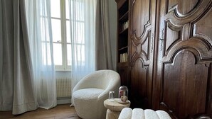 Ein Zimmer mit Holzboden, einem weißen Sessel, einem runden Hocker, einem Holzregal und einem Fenster mit durchscheinenden Vorhängen.
