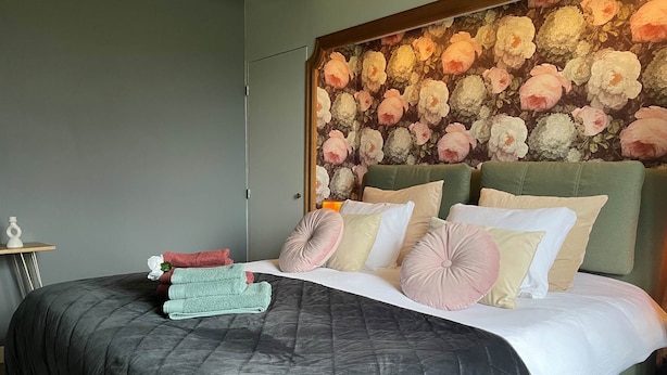 Une chambre à coucher avec une couvre-lit matelassée de couleur sombre, une tête de lit verte et un papier peint à motifs floraux. On peut voir plusieurs oreillers et des serviettes pliées sur le lit.