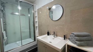 Une salle de bain moderne avec une douche à parois de verre, un lavabo blanc avec un miroir et des serviettes soigneusement pliées sur une étagère.