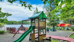Un parque infantil con tobogán, casita de juegos y zona de picnic junto a un lago.