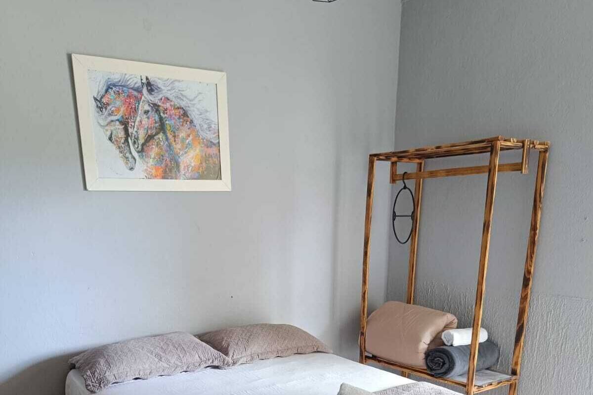 Ein ordentlich bezogenes Bett mit einer braunen Tagesdecke und Kissen, ein hölzerner Stuhl mit Sitzkissen, ein kleiner Nachttisch und ein Wandbild eines Pferdes.