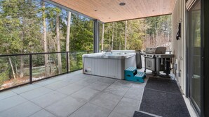 Um terraço moderno ao ar livre com banheira de hidromassagem, churrasqueira e vista para uma área arborizada.