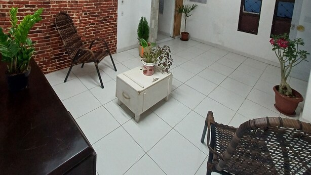 Une pièce spacieuse avec un mur en briques, un sol carrelé de carreaux blancs, une table en bois, deux chaises en osier, une armoire blanche et plusieurs plantes en pot.