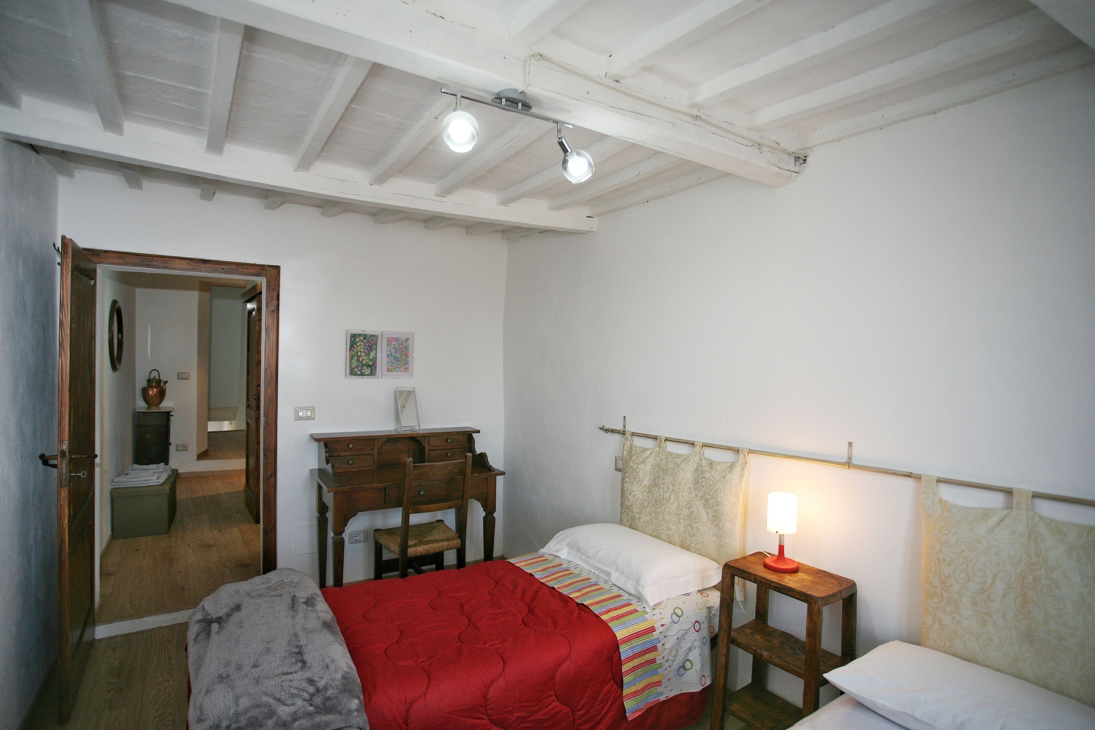 A bedroom with a bed, a desk, a chair, a small table with a lamp, and a door leading to another room.