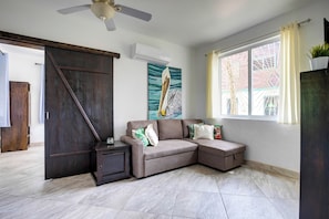 Ein Wohnzimmer mit Ecksofa, Deckenventilator, großem Fenster mit Vorhängen und einem Gemälde an der Wand.