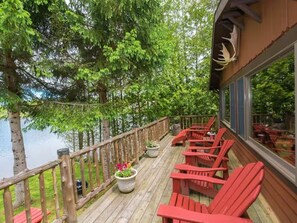 Une terrasse en bois avec des fauteuils Adirondack rouges, donnant sur un lac et entourée d’arbres.