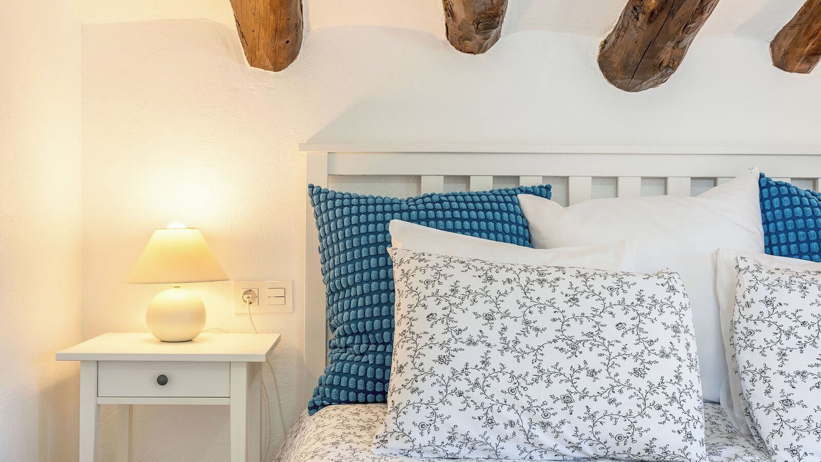 A bedroom with a white bed, blue patterned pillows, a white nightstand with a lamp, and exposed wooden beams on the ceiling.