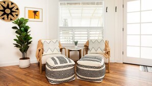Une pièce chaleureuse avec un plafond en bois, une grande fenêtre avec des stores, deux fauteuils ornés de coussins à motifs, une petite table et une plante en pot.