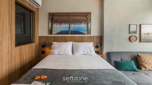 Egy modern szállodaszoba, amelyben található egy ágy, egy kanapé, egy nagy ablak, egy légkondicionáló és egy falra szerelt műalkotás.