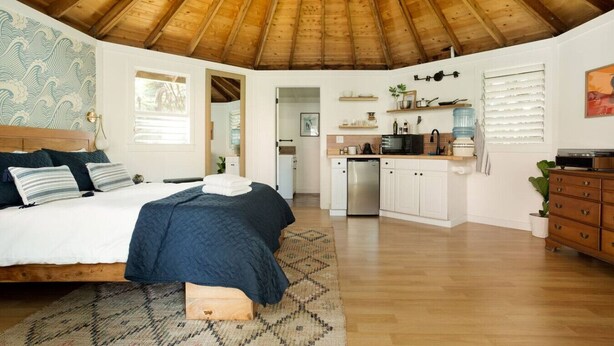 A bedroom with a large bed, a wooden ceiling, and a view into the kitchen area.