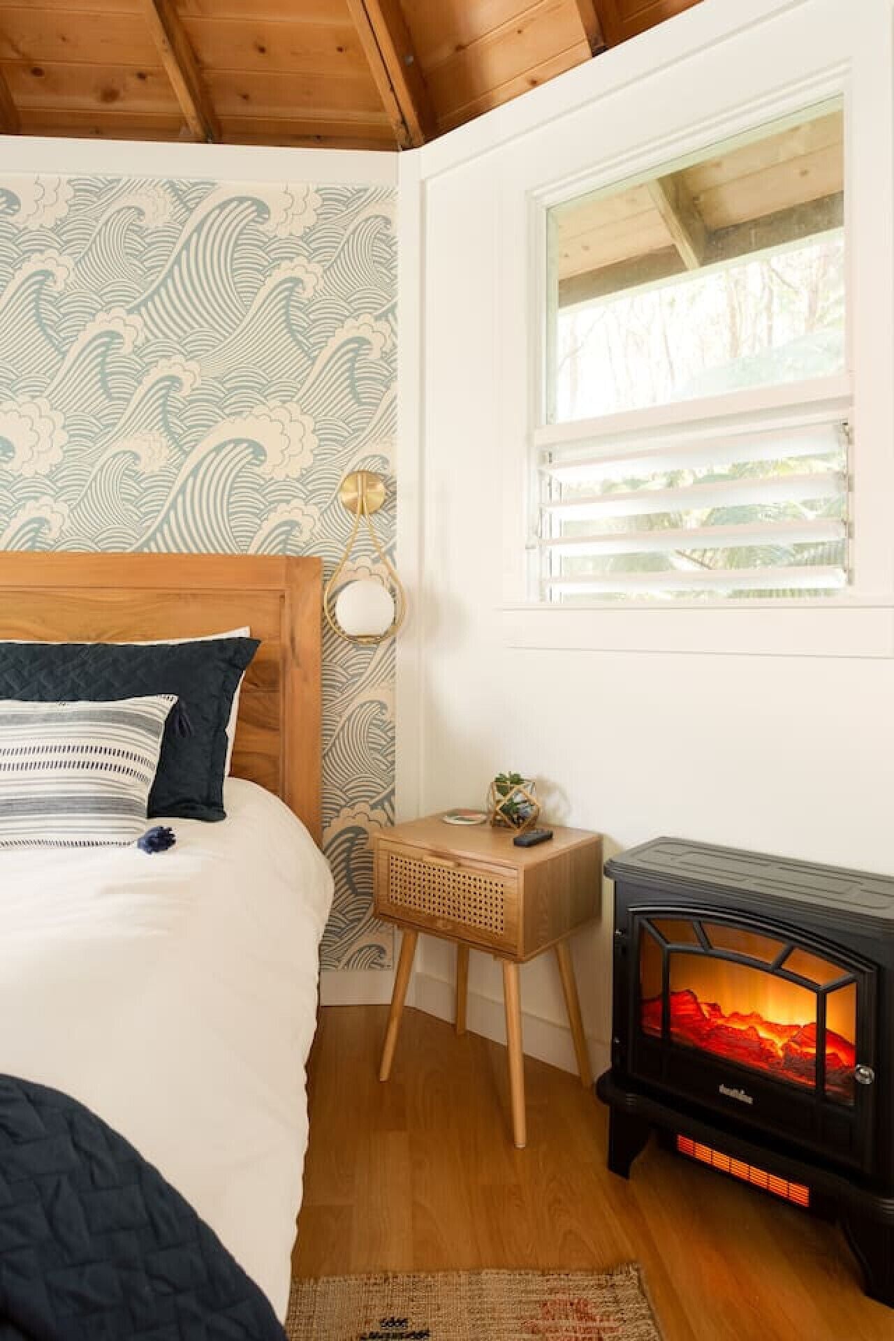 Une chambre à coucher avec un lit, une petite table de chevet ornée d’une plante, une lampe fixée au mur et un poêle à bois.