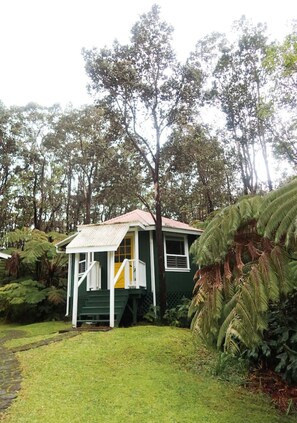 A small, single-story house with a green exterior and a red roof, surrounded by lush greenery and trees. - Ohia Cottage Volcano Hawaii (Volcano)