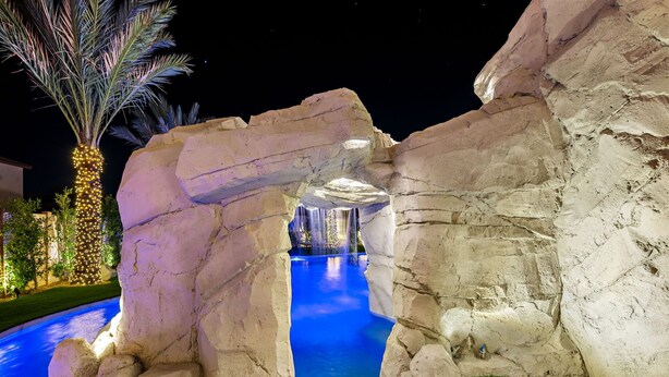 A nighttime pool area with a rock archway, illuminated water, and a palm tree. - Isle of Palms: Sleeps 27, Lazy River & Waterslide (Indio)