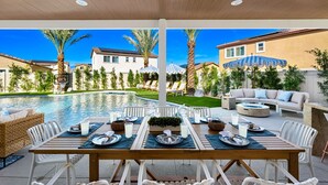 An outdoor dining area with a pool view, white furniture, and a well-maintained lawn. - Isle of Palms: Sleeps 27, Lazy River & Waterslide (Indio)