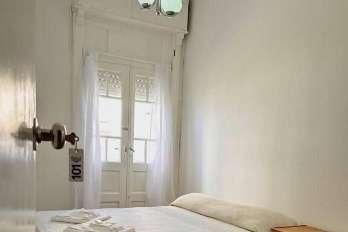Una habitación con cama, mesita de noche de madera, puerta con tarjeta de acceso y ventana con cortinas.