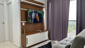 A modern living room with a flat-screen TV mounted on a wooden wall, a sofa, and a view of greenery outside. - Apto. 2 quartos Vista Rio Negro P. Negra (Manaus)