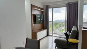 A modern living room with a sofa, a TV mounted on a wooden panel, a dining table with chairs, and a balcony with a view. - Apto. 2 quartos Vista Rio Negro P. Negra (Manaus)