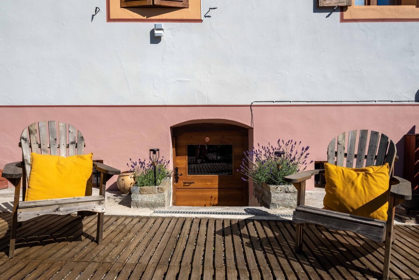 Une terrasse en bois avec deux fauteuils Adirondack, une porte en bois et un petit jardin agrémenté de plantes de lavande.