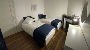 Et soveværelse med to senge, en hvid dør, et inspirerende citat i ramme på væggen, et lille bord med en lampe og en pejs.