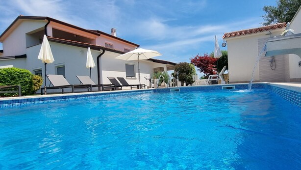 Ein Swimmingpool mit Liegestühlen und Sonnenschirmen, umgeben von Gebäuden mit einem Ziegeldach und weißen Wänden.