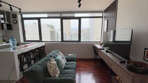 Una sala de estar moderna con un sofá verde, una zona de comedor con barra, un ventanal con vistas a la ciudad y un televisor de pantalla plana.