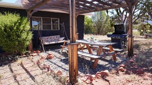 Una pérgola de madera con mesa de picnic y zona de barbacoa, rodeada de grava y plantas.