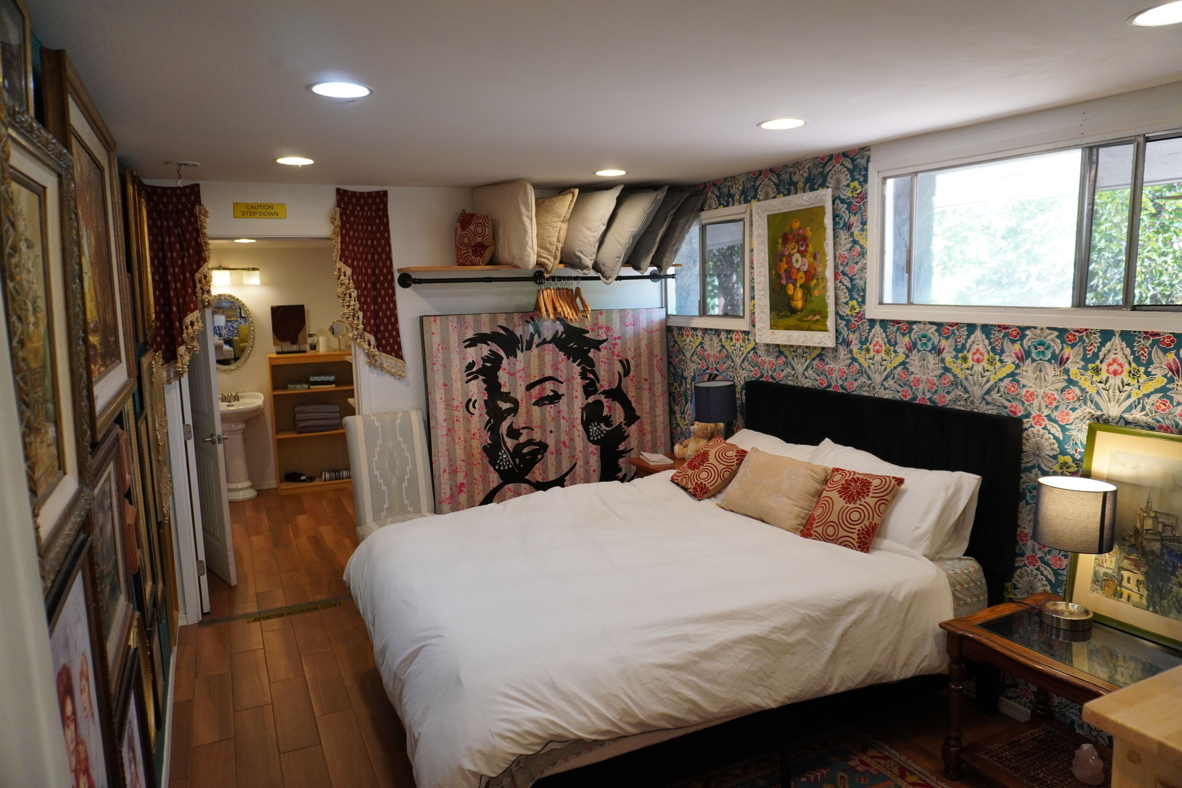 Une chambre à coucher avec un grand lit, une table de chevet, un miroir, une fenêtre et un mur recouvert d’un papier peint à motifs floraux.