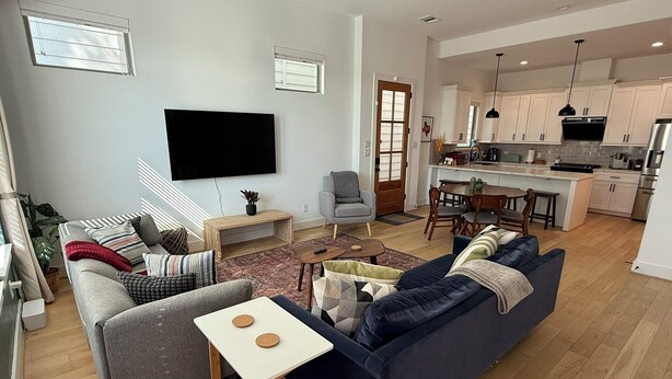 A modern living room with a sofa, a coffee table, and a TV. There is a kitchen in the background with cabinets and a dining area. - Parkside Central Retreat (Houston)