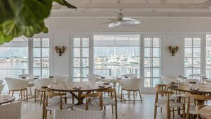 Un comedor con mesas y sillas redondas, ventanas amplias con vista al puerto deportivo y ventiladores de techo.