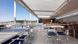 Un área de comedor al aire libre techada con mesas y sillas, con vista a un puerto deportivo con barcos.