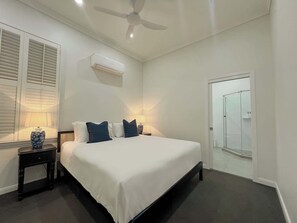A bedroom with a bed, bedside lamps, a ceiling fan, an air conditioning unit, and a glass shower door.