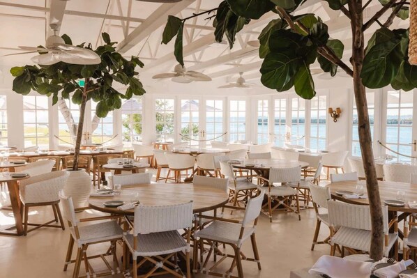 A spacious dining area with round tables and white chairs, large windows with a view of the sea, and hanging plants.