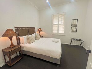 A neatly made bed with white linens and two pillows, flanked by two bedside tables with lamps, a window with blinds, and a radiator.