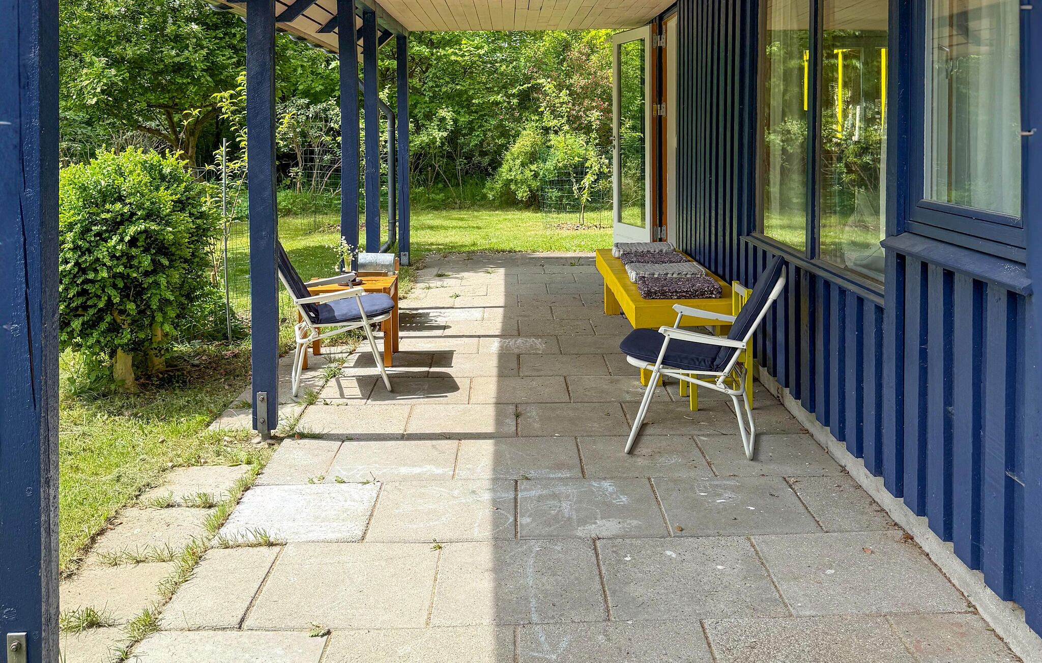 A covered patio with two yellow tables and two chairs, surrounded by a blue building with large windows.