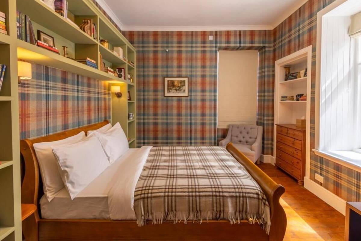 A bedroom with a wooden bed, green bookshelves, plaid wallpaper, and a window with curtains.