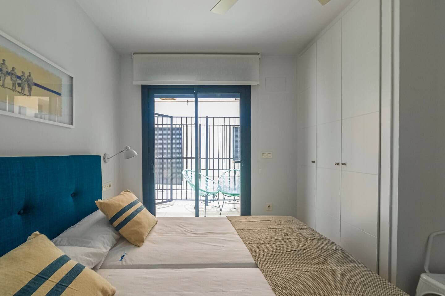 A bedroom with a bed, a desk, and a chair. There is a window with blinds and a door leading to a balcony.