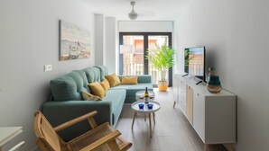 A modern living room with a sofa, a wooden chair, a small round table with drinks, a TV on a cabinet, and a large window with a view of buildings. - Costa ap. 2nd floor shared heated pool (Alicante/Alacant)