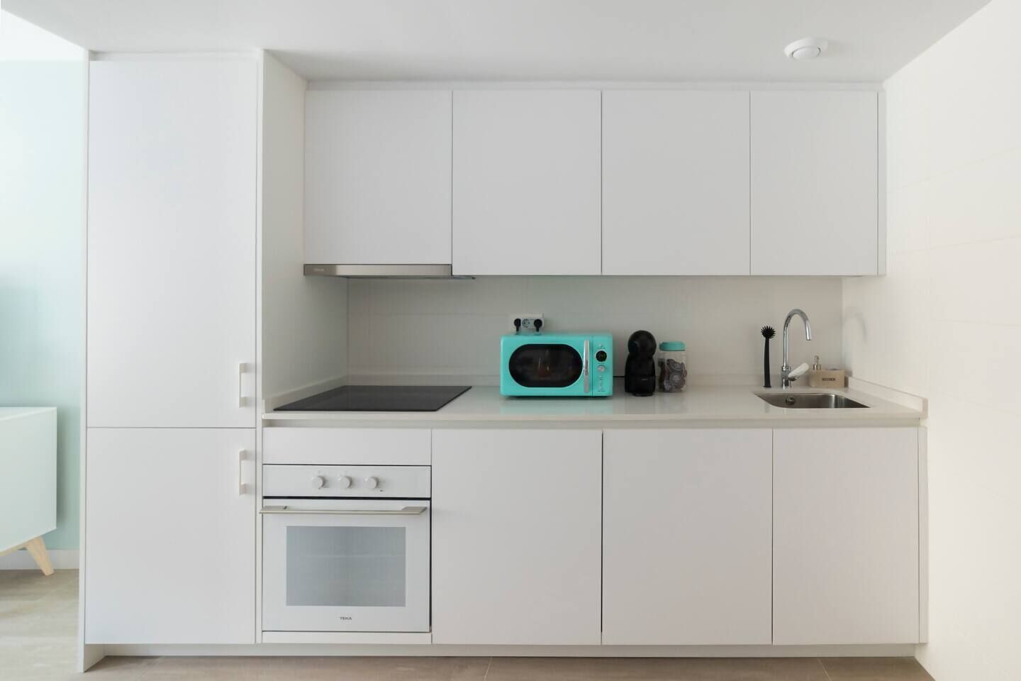 A modern kitchen with white cabinetry, a black stovetop, a teal microwave, and a stainless steel sink.