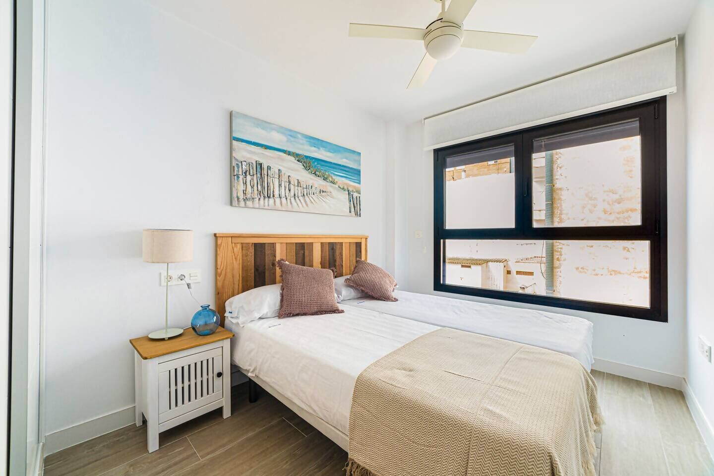 Une chambre à coucher comprenant un lit, une table de chevet, une lampe, un ventilateur de plafond, une fenêtre et un tableau accroché au mur.