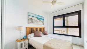 A bedroom with a bed, a nightstand, a lamp, a ceiling fan, a window, and a painting on the wall. - Ap. Marino 2nd floor shared heated swimming pool (Alicante/Alacant)