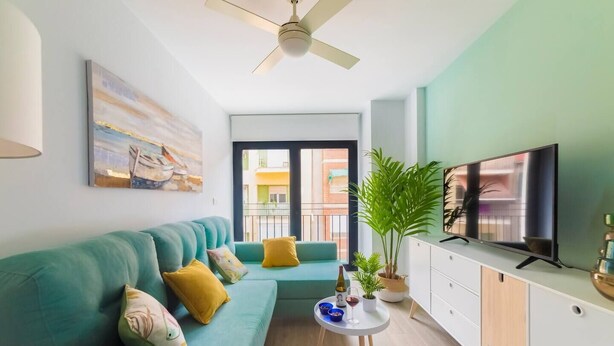 A modern living room with a teal sofa, a flat-screen TV, a ceiling fan, and a large window with a view of buildings. - Ap. Marino 2nd floor shared heated swimming pool (Alicante/Alacant)