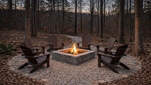 Een vuurkuil met vier Adirondack-stoelen eromheen, gelegen op een grindpad omringd door bomen.