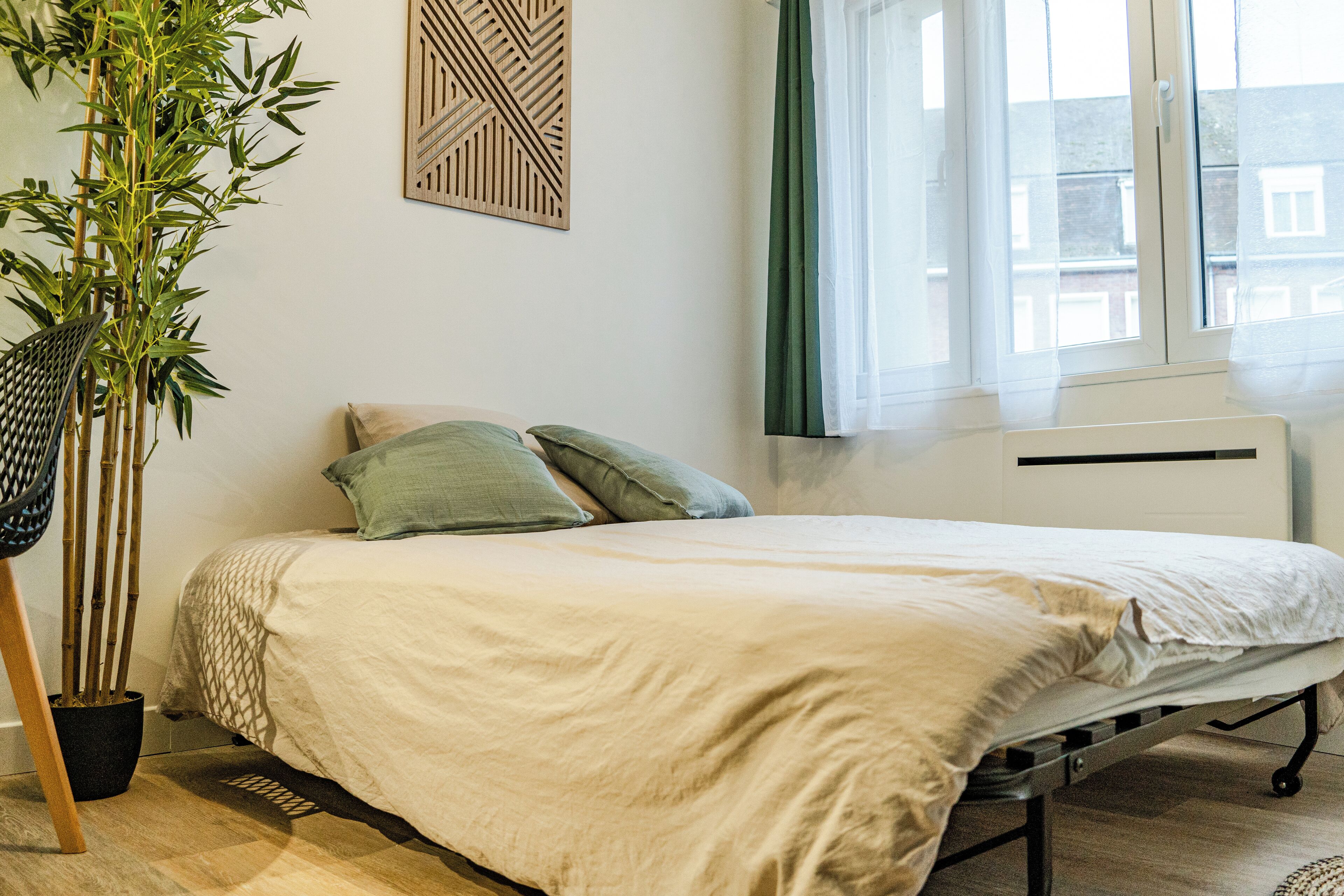 Ein Schlafzimmer mit einem Bett, einem Topf mit Pflanze, einem Fenster mit Vorhängen und einer Heizung.
