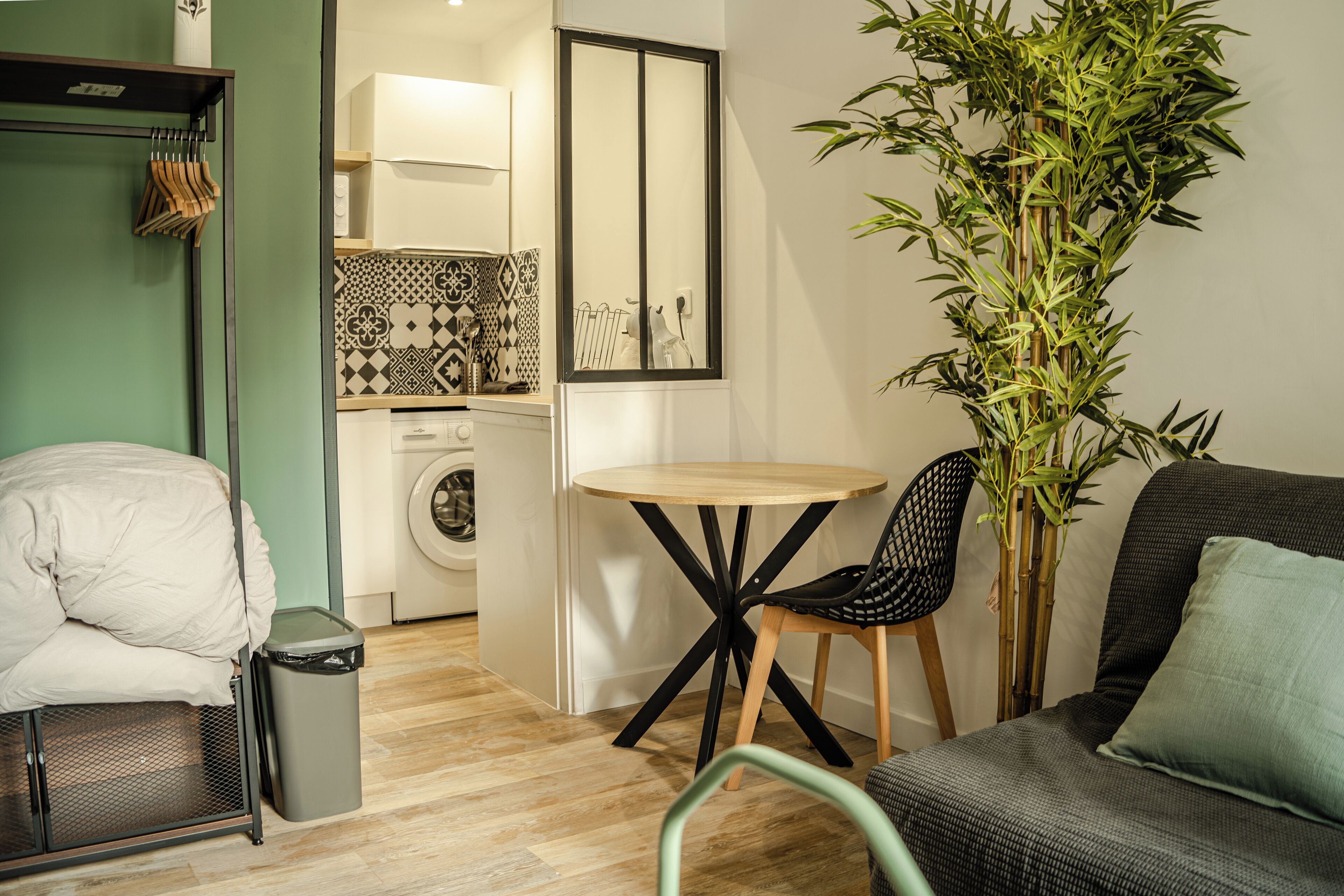 A compact living space with a bed, a small table, a chair, a washing machine, and a large plant.
