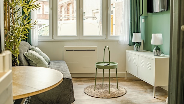 A room with a sofa, a small table, a radiator, a TV mounted on the wall, and a view of the street outside. - CosyEmerald (Cambrai)